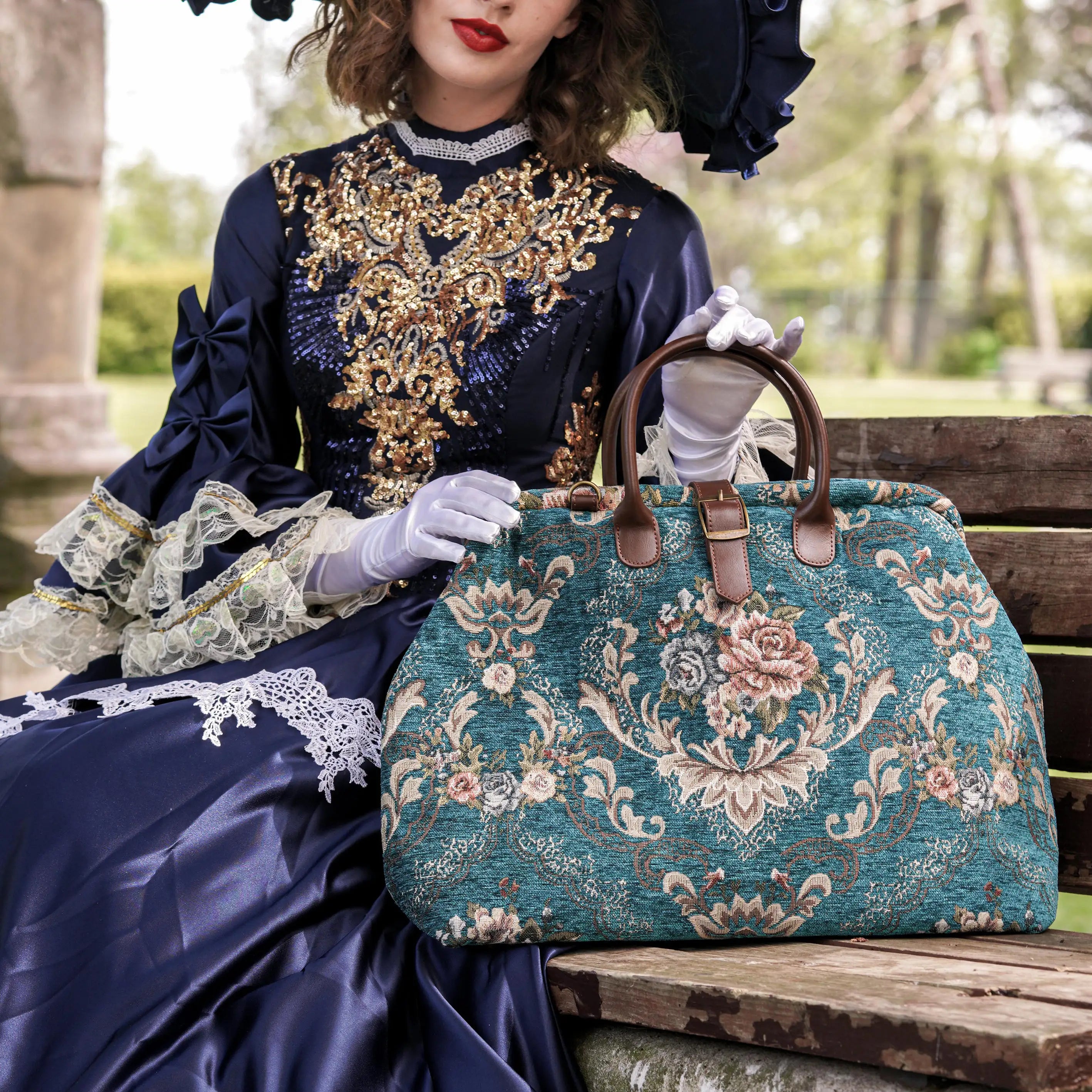 Elegant Victorian-inspired tapestry travel bag with floral chenille pattern, leather handles, and vintage styling, held by a model in a navy embroidered dress.
