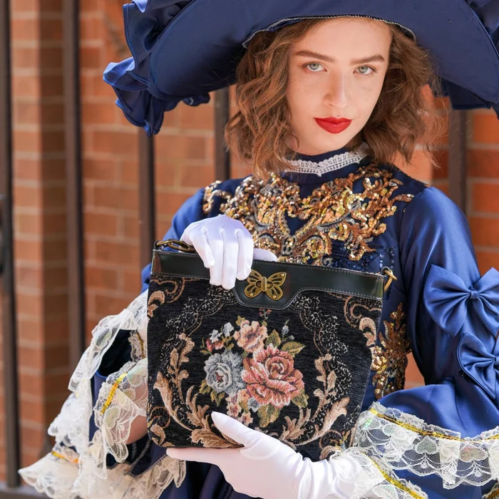Woman in elegant Victorian-inspired blue dress holding a black floral tapestry carpet crossbody bag with butterfly clasp and leather trim.