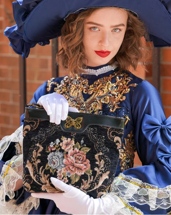 Woman in elegant Victorian-inspired blue dress holding a black floral tapestry carpet crossbody bag with butterfly clasp and leather trim.