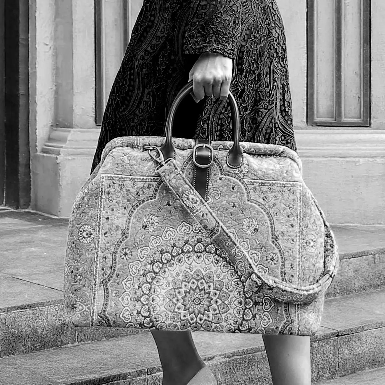 Victorian-inspired carpet weekender bag with intricate tapestry pattern, leather handles, and vintage brass closure, carried by a model in a black lace dress.