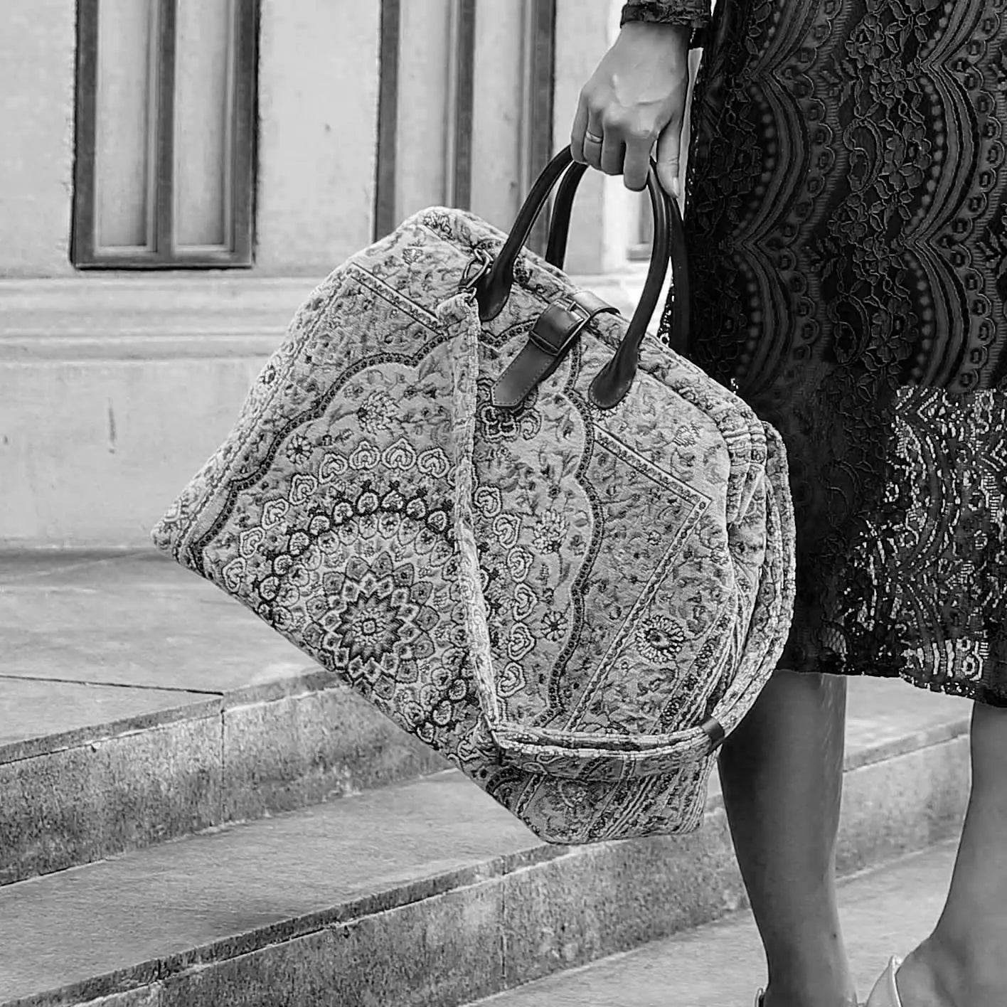 Victorian-inspired carpet weekender bag with intricate tapestry pattern, leather handles, and vintage brass closure, carried by a model in a black lace dress.