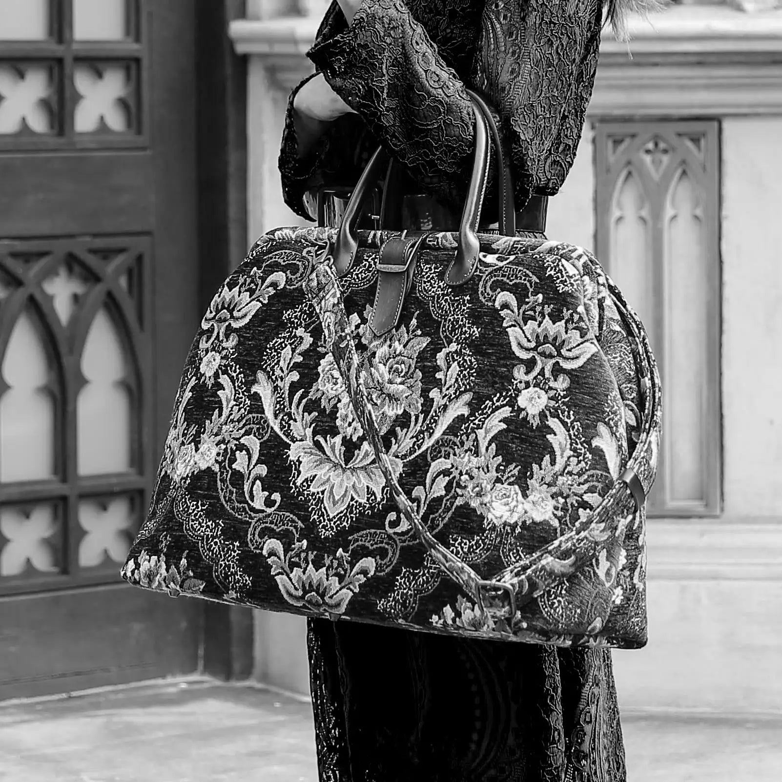 Elegant black floral tapestry travel bag with leather handles and detachable strap, styled with a model in a gothic Victorian lace dress.