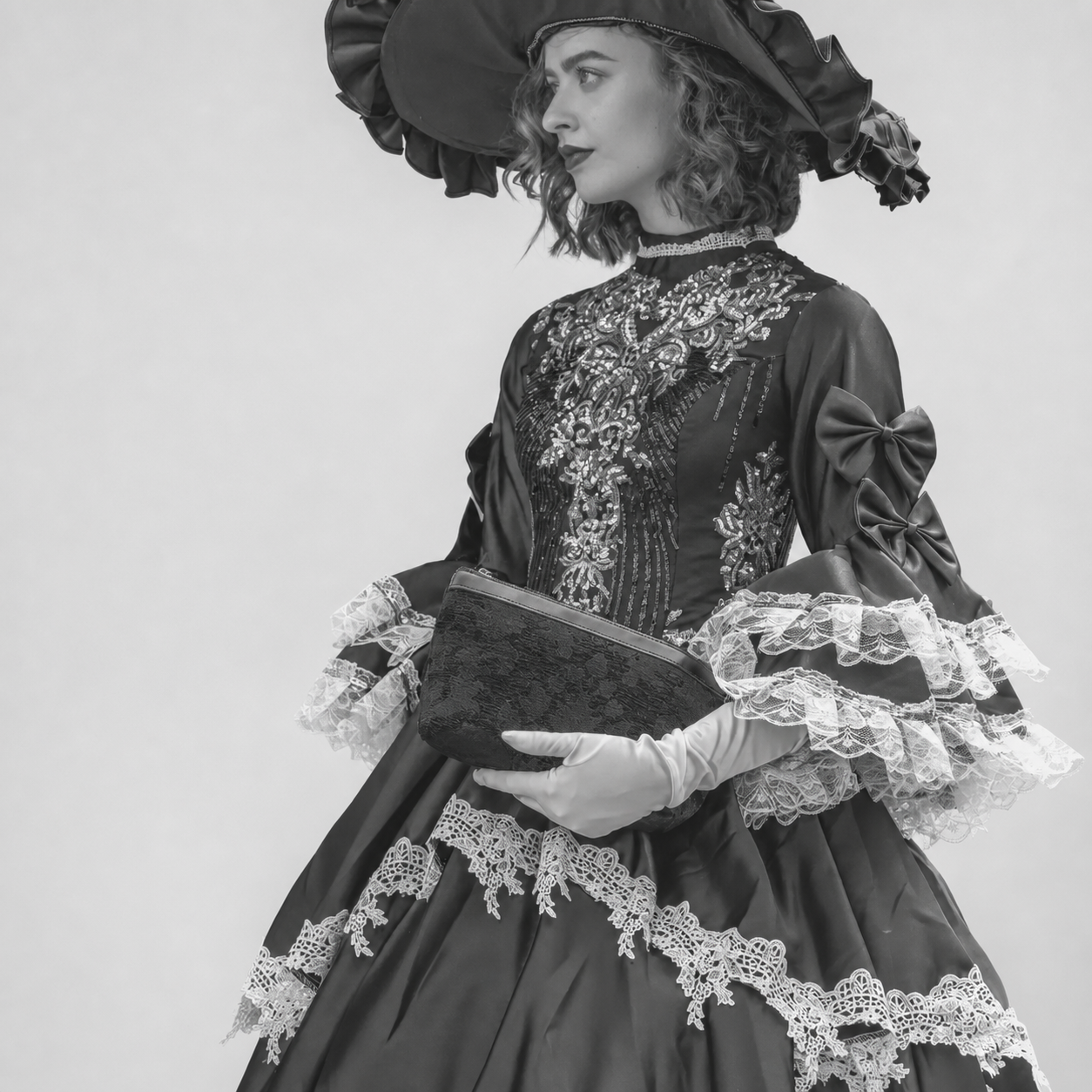 A woman wearing an ornate Victorian-style black dress with intricate lace embroidery, ruffled sleeves, lace trim, a large bow, and a matching wide-brimmed hat, posed against a plain light background while holding a dark clutch.