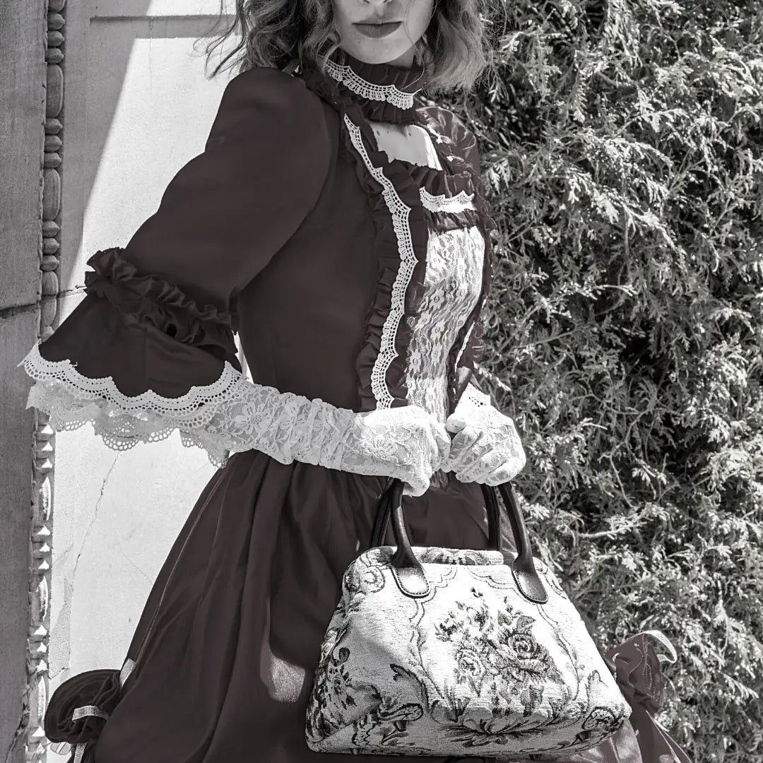 Victorian-style floral carpet handbag with leather handles, carried by a woman wearing a vintage-inspired lace dress and gloves.