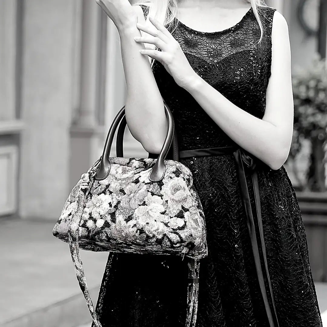 Floral chenille carpet handbag with leather handles and detachable shoulder strap, carried by a woman in a black lace dress.