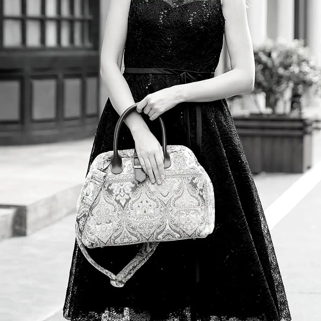 Woman wearing a black sequined dress carrying a vintage-inspired carpet handbag with leather handles and shoulder strap in an elegant city setting.