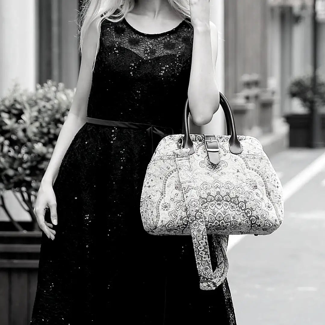 Woman wearing a black sequined dress carrying a vintage-inspired carpet handbag with leather handles and shoulder strap in an elegant city setting.