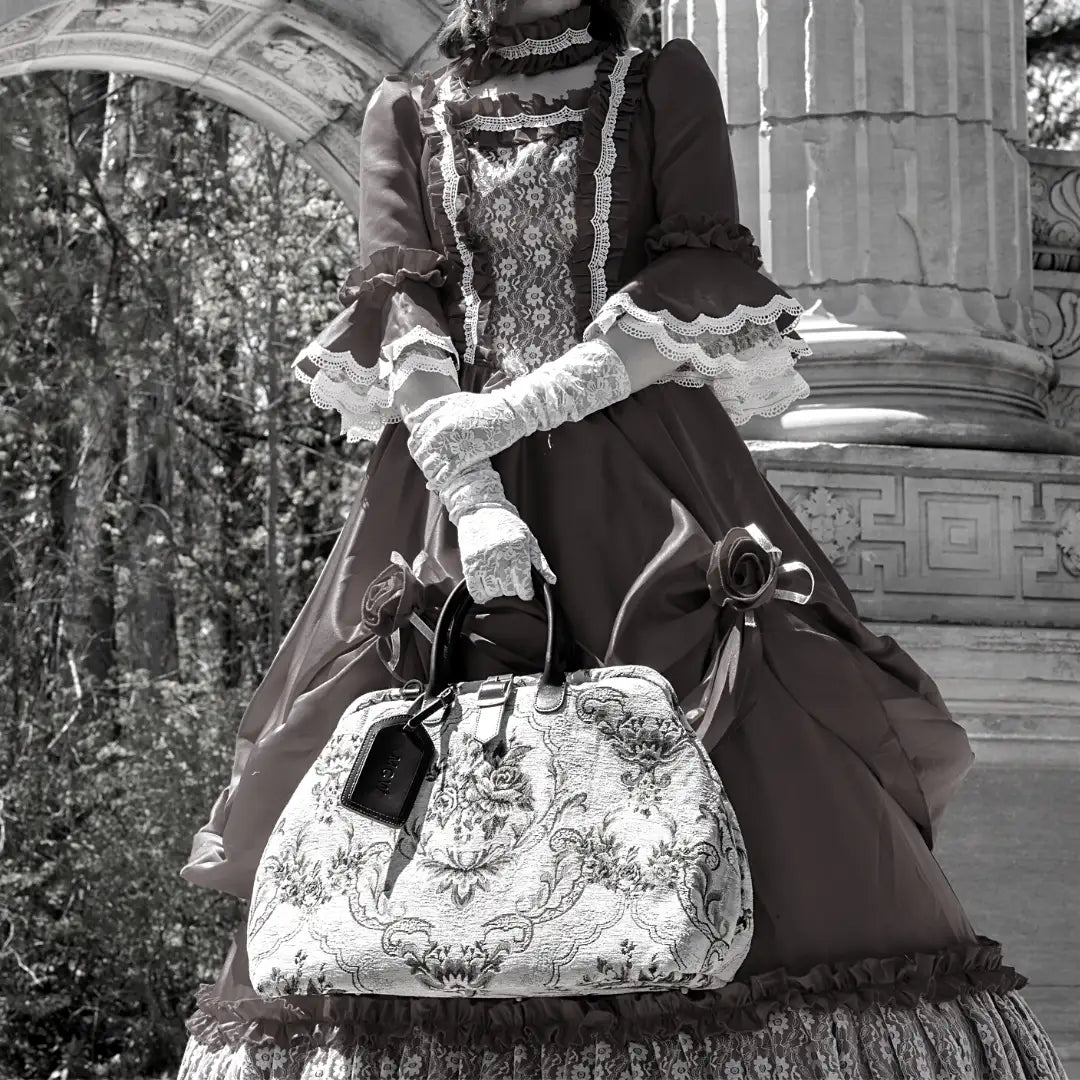 Victorian-inspired floral tapestry travel bag with leather handles and luggage tag, styled with a model in an elegant lace-trimmed historical gown.