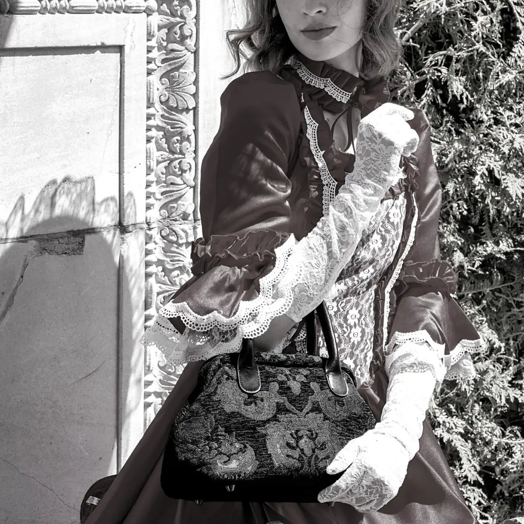 Victorian-style floral carpet handbag with leather handles, carried by a woman wearing a vintage-inspired lace dress and gloves.