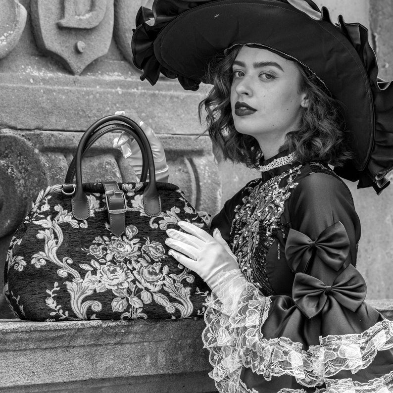 Woman in Victorian-inspired black lace dress and wide-brim hat holding a vintage floral carpet bag beside an ornate stone balcony in an elegant gothic setting.