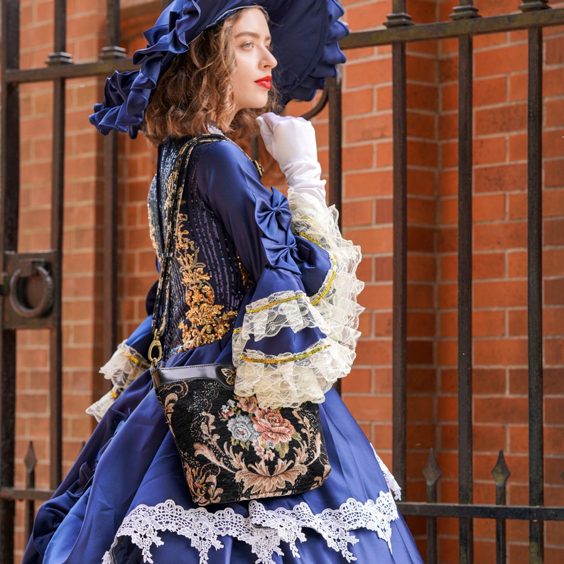 Woman in a Victorian-inspired navy dress with lace details and a wide-brimmed hat, carrying a floral tapestry shoulder bag with a butterfly clasp while posing beside an iron gate.