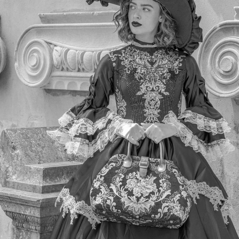 Woman in Victorian-inspired black lace dress and wide-brim hat holding a vintage floral carpet bag beside an ornate stone balcony in an elegant gothic setting.
