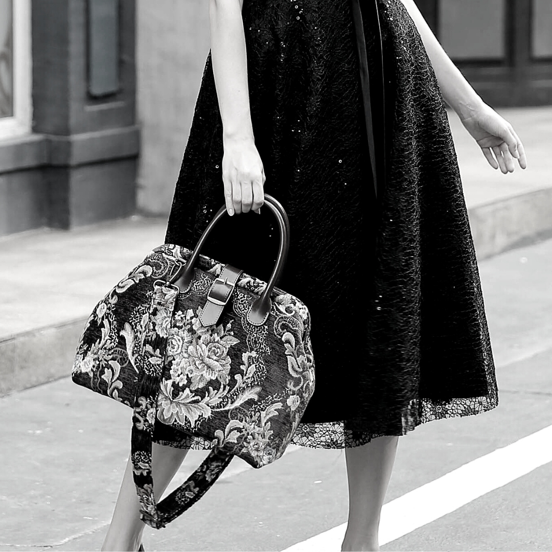 Woman wearing a black sequined dress carrying a vintage-inspired carpet handbag with leather handles and shoulder strap in an elegant city setting.