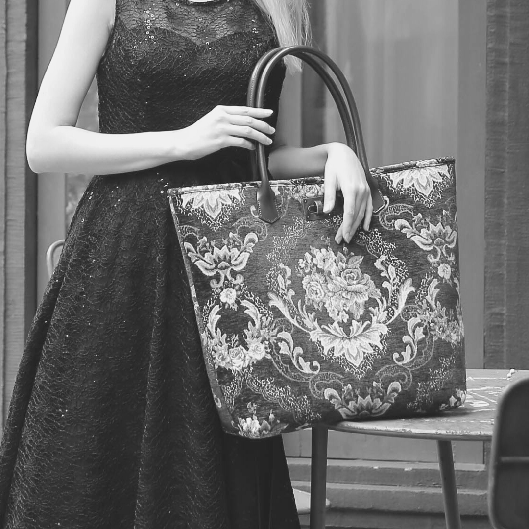 Large floral tapestry tote bag with brown leather shoulder straps, featuring an ornate Victorian-inspired rose pattern. Shown carried by a model in a black dress, highlighting its spacious design and elegant vintage style.