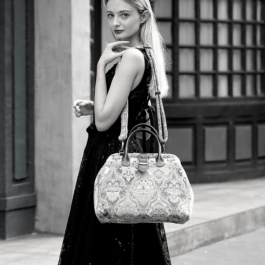Woman wearing a black sequined dress carrying a vintage-inspired carpet handbag with leather handles and shoulder strap in an elegant city setting.
