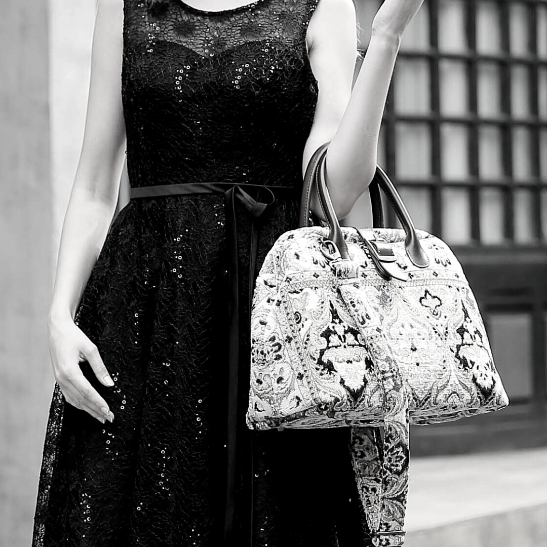 Woman wearing a black sequined dress carrying a vintage-inspired carpet handbag with leather handles and shoulder strap in an elegant city setting.