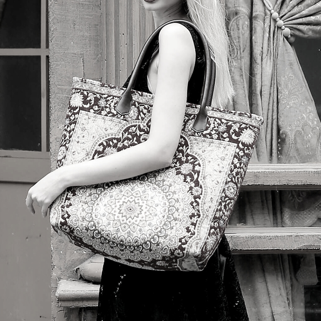 Large tapestry tote bag with an ornate floral medallion pattern and dark leather shoulder straps, carried by a model in front of a vintage stone building.