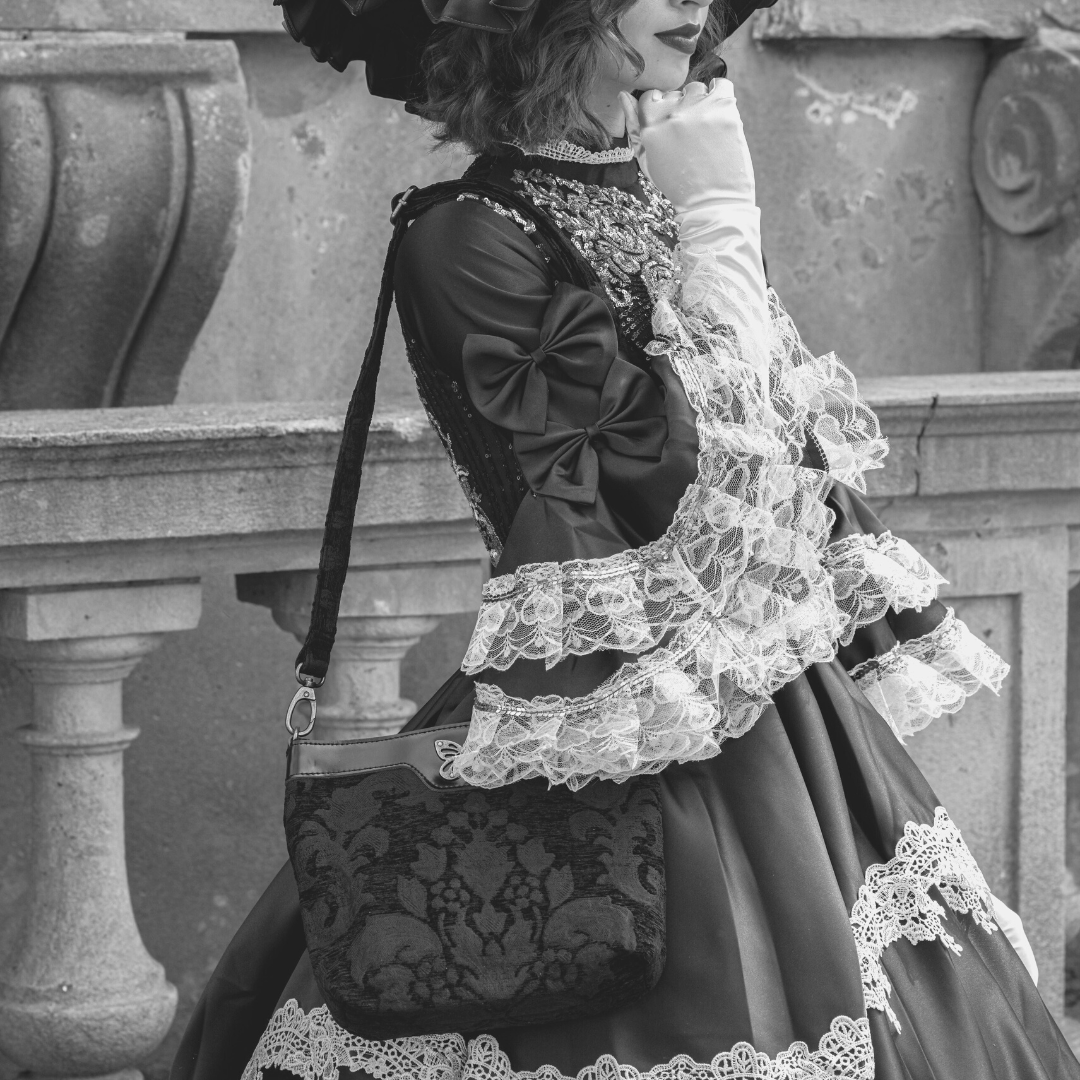 Woman in a black Victorian-inspired dress with lace cuffs and a wide-brimmed hat, holding a floral tapestry shoulder bag with a butterfly clasp and leather trim.