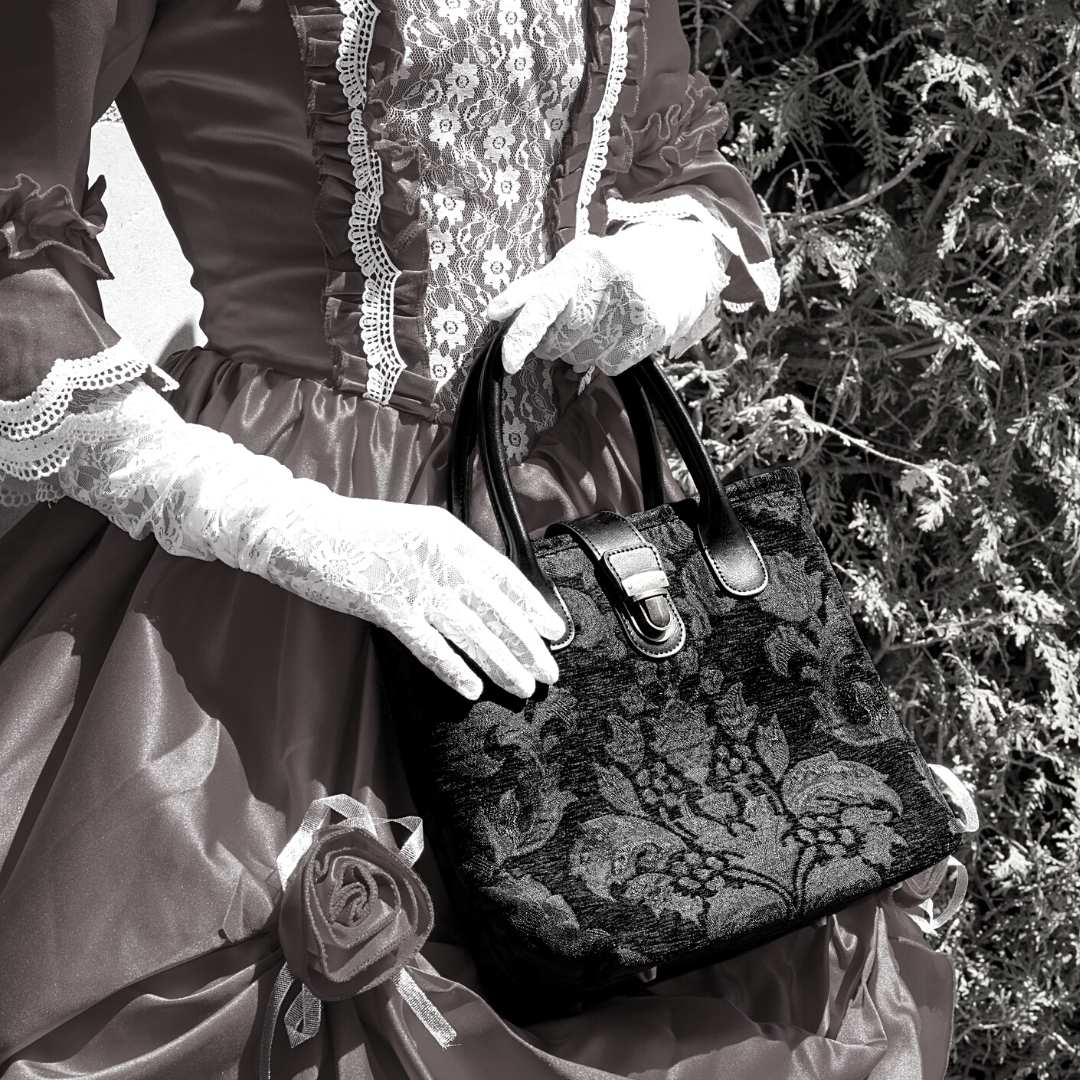 Victorian-style woman in a lace-trimmed dress and gloves holding a black damask tapestry handbag with leather handles and clasp closure.