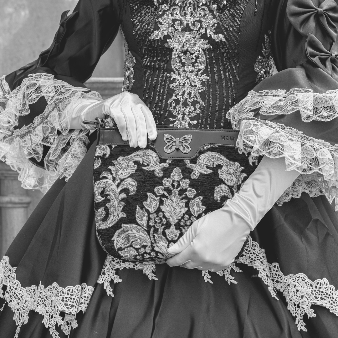 Close-up of a woman in a Victorian-inspired black dress with lace cuffs and white gloves, holding a floral tapestry shoulder bag with a butterfly clasp and leather trim.
