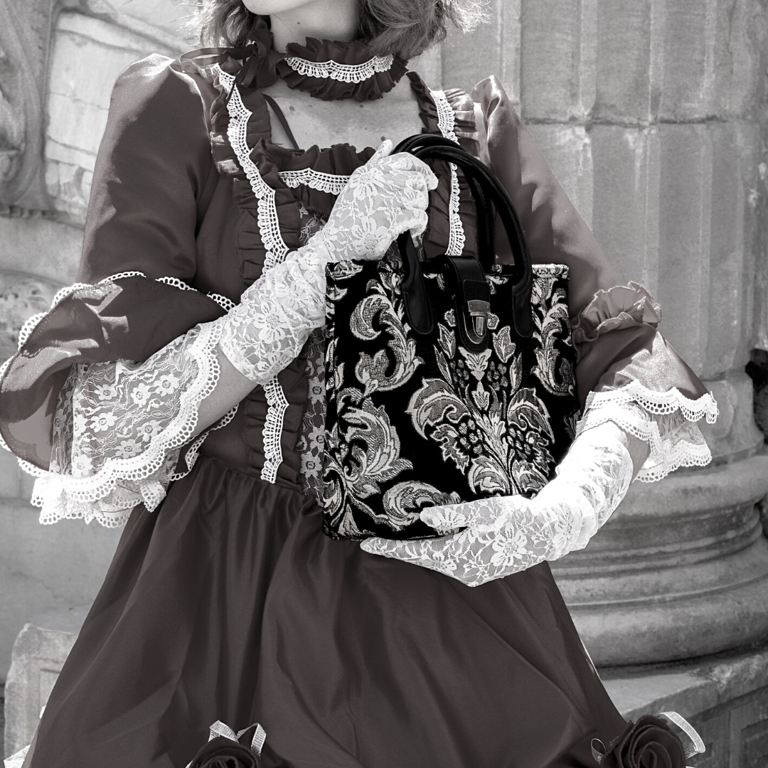 Victorian-style woman in a lace-trimmed dress and gloves holding a black damask tapestry handbag with leather handles and clasp closure.
