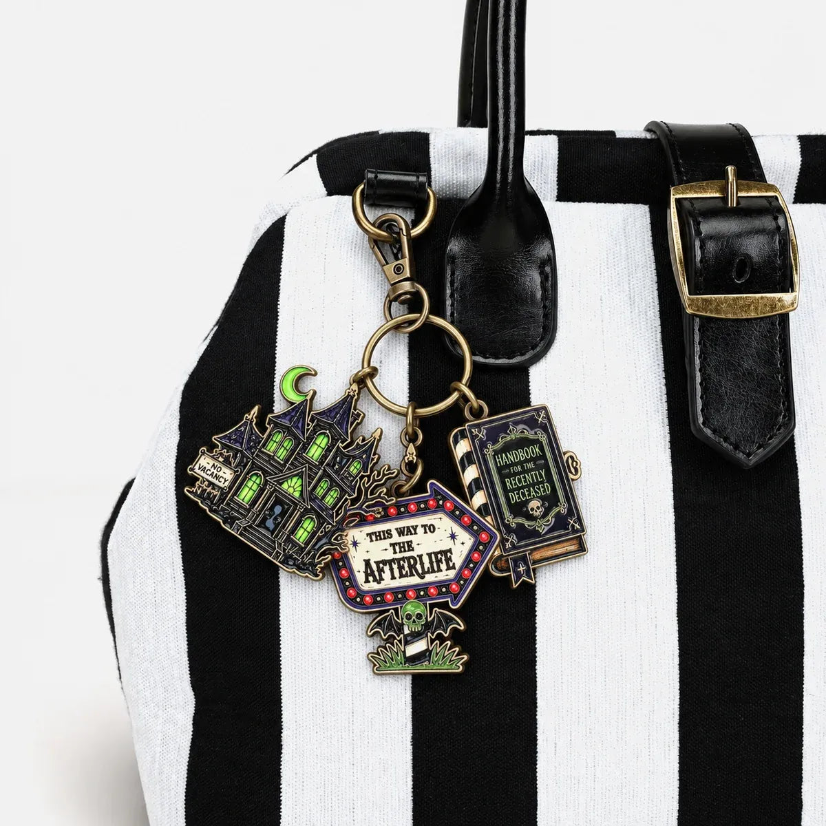 A close-up of a black and white striped tote bag with black leather handles and a gold-tone buckle, featuring three decorative keychain charms shaped like a haunted house, a book, and a sign reading 'THE WAY TO THE AFTERLIFE'.