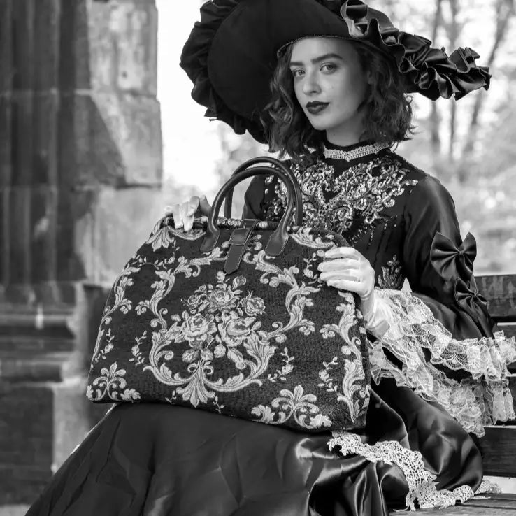 Elegant floral tapestry travel bag with leather handles, styled with a model in a black Victorian-inspired dress and dramatic wide-brim hat.