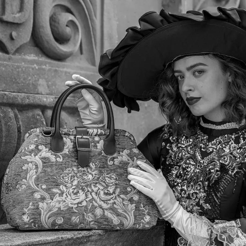 Woman in Victorian-inspired black lace dress and wide-brim hat holding a vintage floral carpet bag beside an ornate stone balcony in an elegant gothic setting.