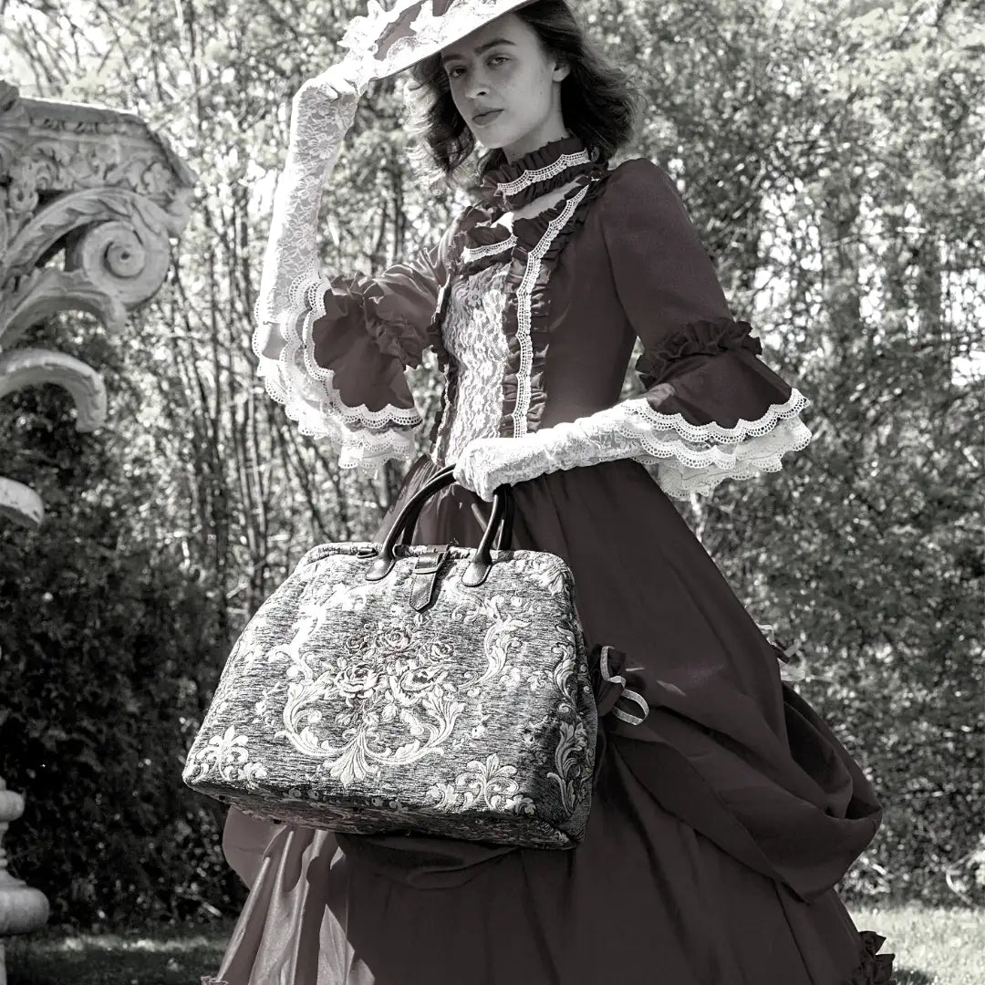 Victorian-inspired floral tapestry travel bag with leather handles, carried by a model in an elegant lace-trimmed historical dress and wide-brim hat.