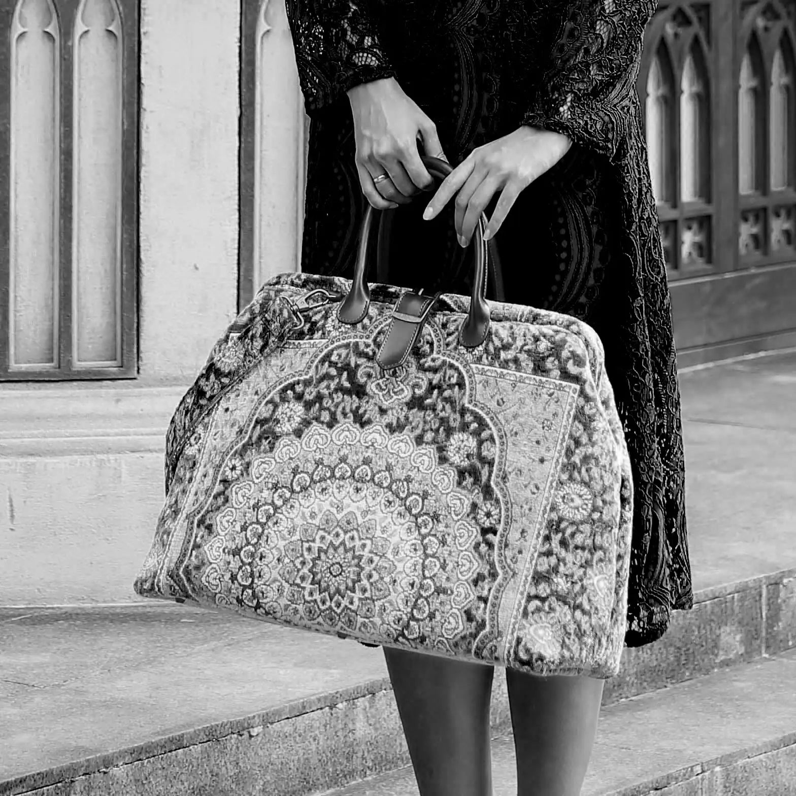 Victorian-inspired carpet weekender bag with intricate tapestry pattern, leather handles, and vintage brass closure, carried by a model in a black lace dress.