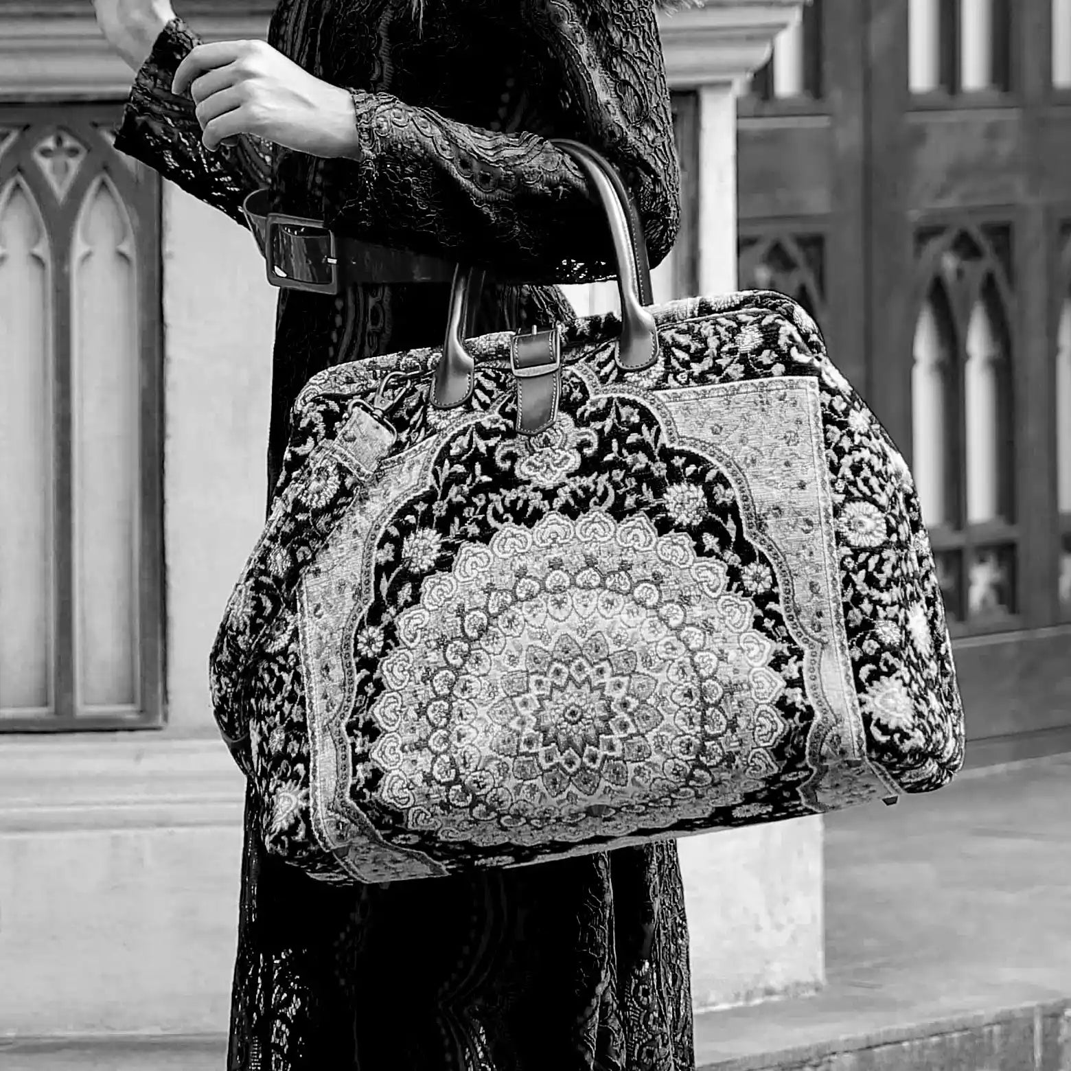 Victorian-inspired carpet weekender bag with intricate tapestry pattern, leather handles, and vintage brass closure, carried by a model in a black lace dress.
