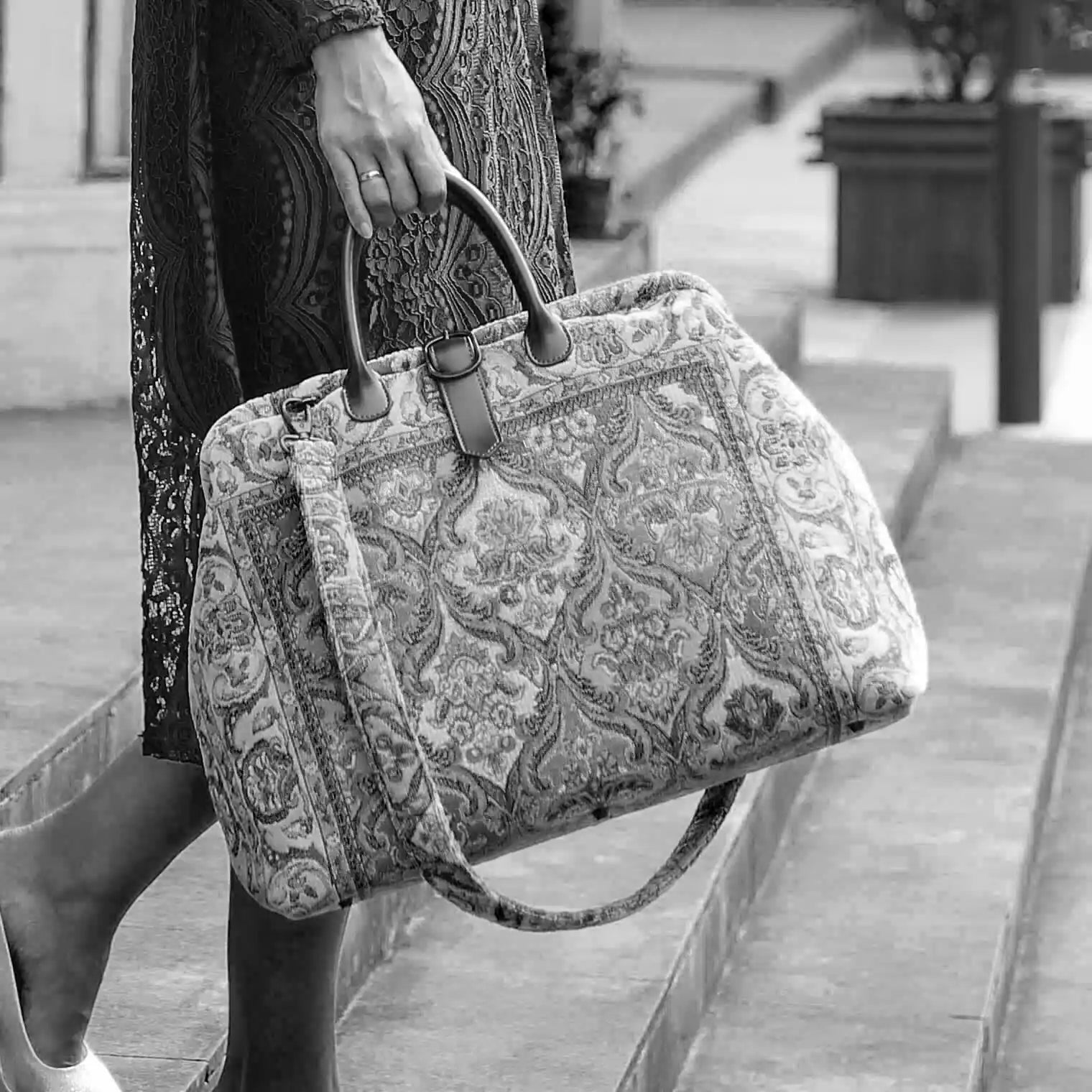 Victorian-inspired carpet weekender bag with intricate tapestry pattern, leather handles, and vintage brass closure, carried by a model in a black lace dress.
