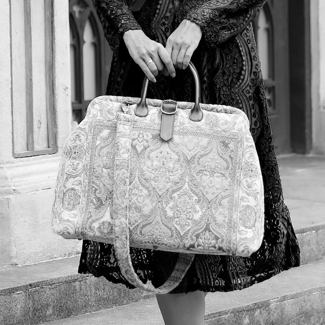 Victorian-inspired carpet weekender bag with intricate tapestry pattern, leather handles, and vintage brass closure, carried by a model in a black lace dress.