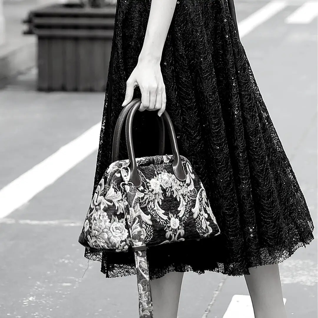 Floral chenille carpet handbag with leather handles and detachable shoulder strap, carried by a woman in a black lace dress.