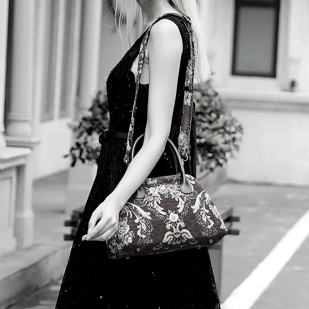Damask chenille carpet handbag with leather handles and detachable shoulder strap, worn over the shoulder by a woman in a black lace dress