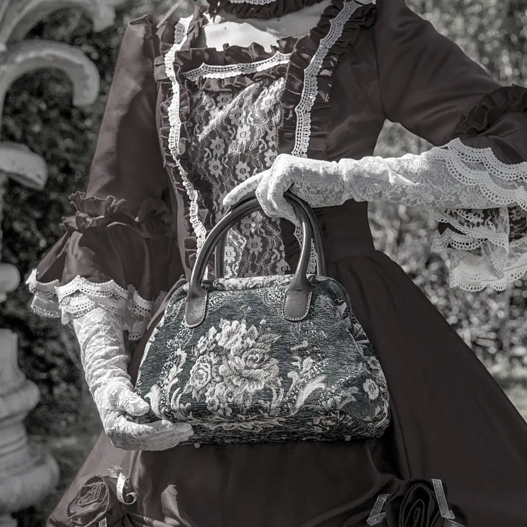 Victorian-style floral carpet handbag with leather handles, carried by a woman wearing a vintage-inspired lace dress and gloves.
