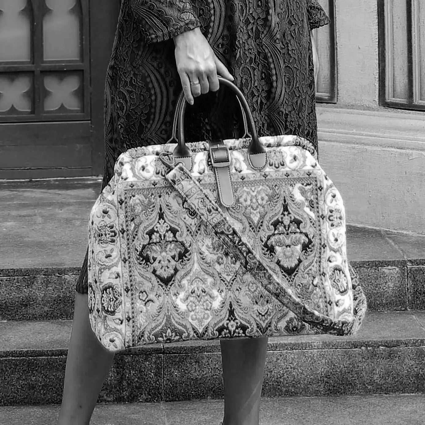 Victorian-inspired carpet weekender bag with intricate tapestry pattern, leather handles, and vintage brass closure, carried by a model in a black lace dress.