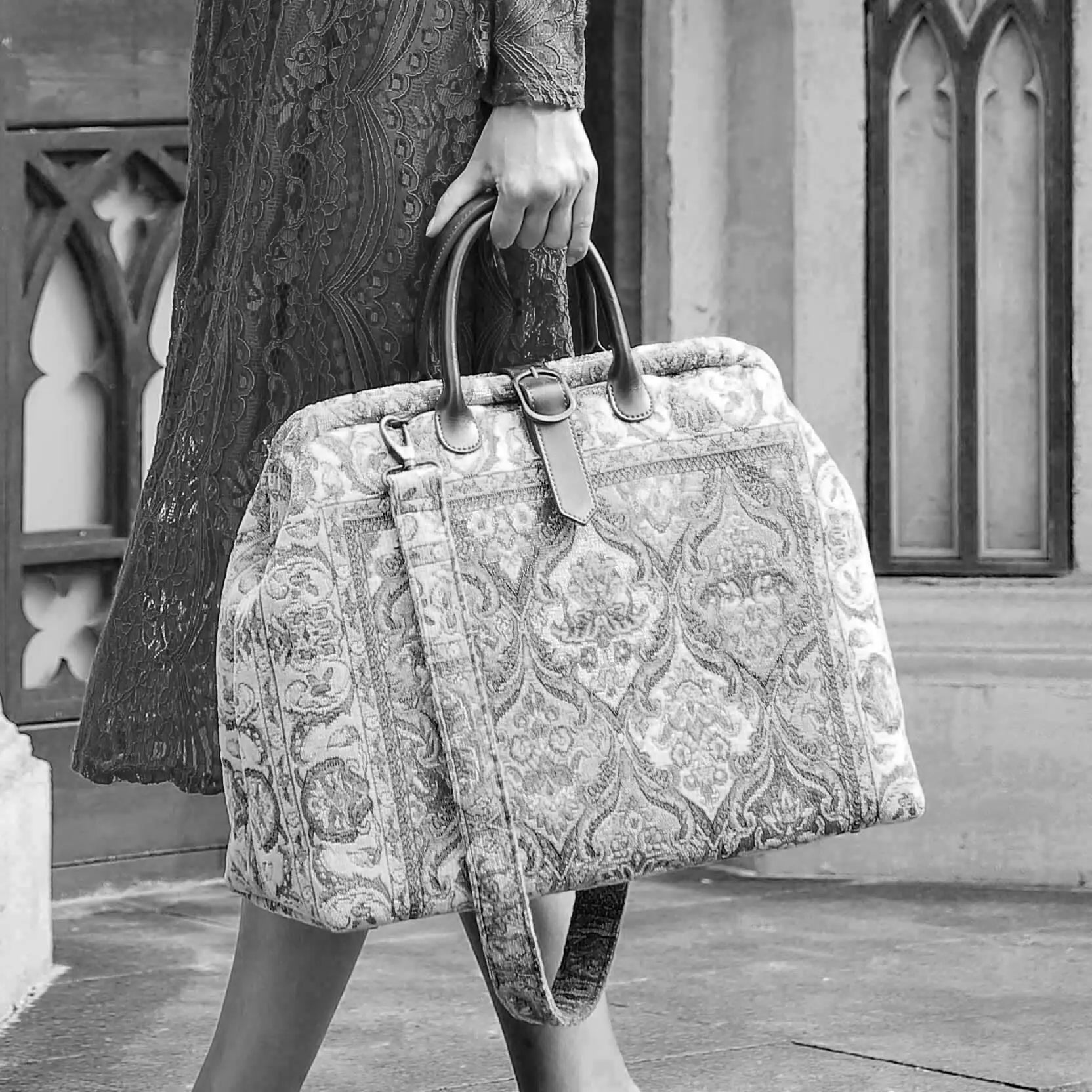 Victorian-inspired carpet weekender bag with intricate tapestry pattern, leather handles, and vintage brass closure, carried by a model in a black lace dress.