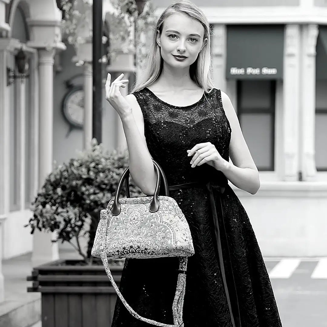 Elegant woman in a black lace dress holding a Victorian-inspired chenille carpet handbag with leather handles and a detachable shoulder strap, standing in a charming European-style street setting.