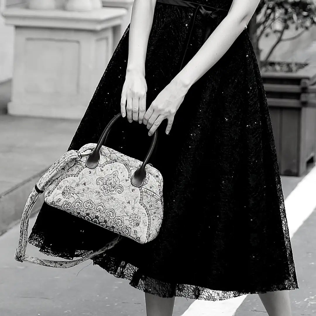 Victorian-inspired chenille carpet handbag with leather handles and detachable shoulder strap, carried by a woman in a black lace dress.