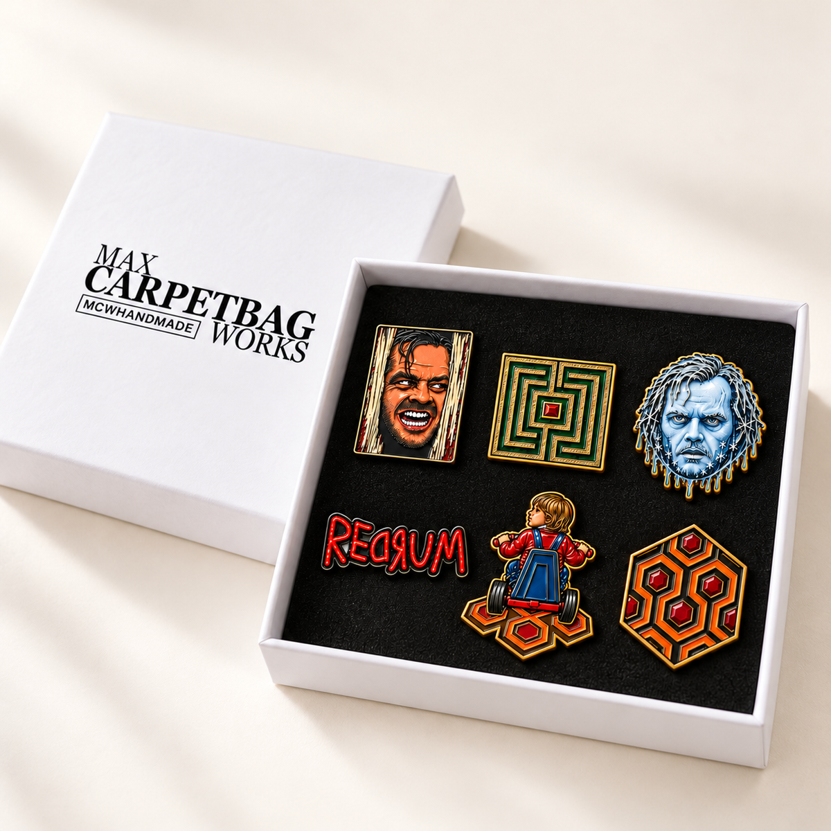 A boxed set of 6 enamel pins displayed in an open white presentation box, with the matching lid placed beside it on a light beige surface. The lid is printed with the brand name 'Max Carpetbag Works' and 'MCWHANDMADE'.
