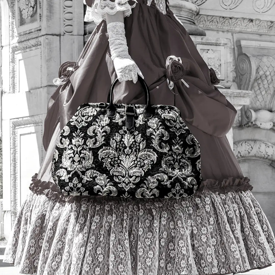 Damask-inspired black chenille travel bag with leather handles and vintage frame styling, carried by a model in an elegant Victorian gown.
