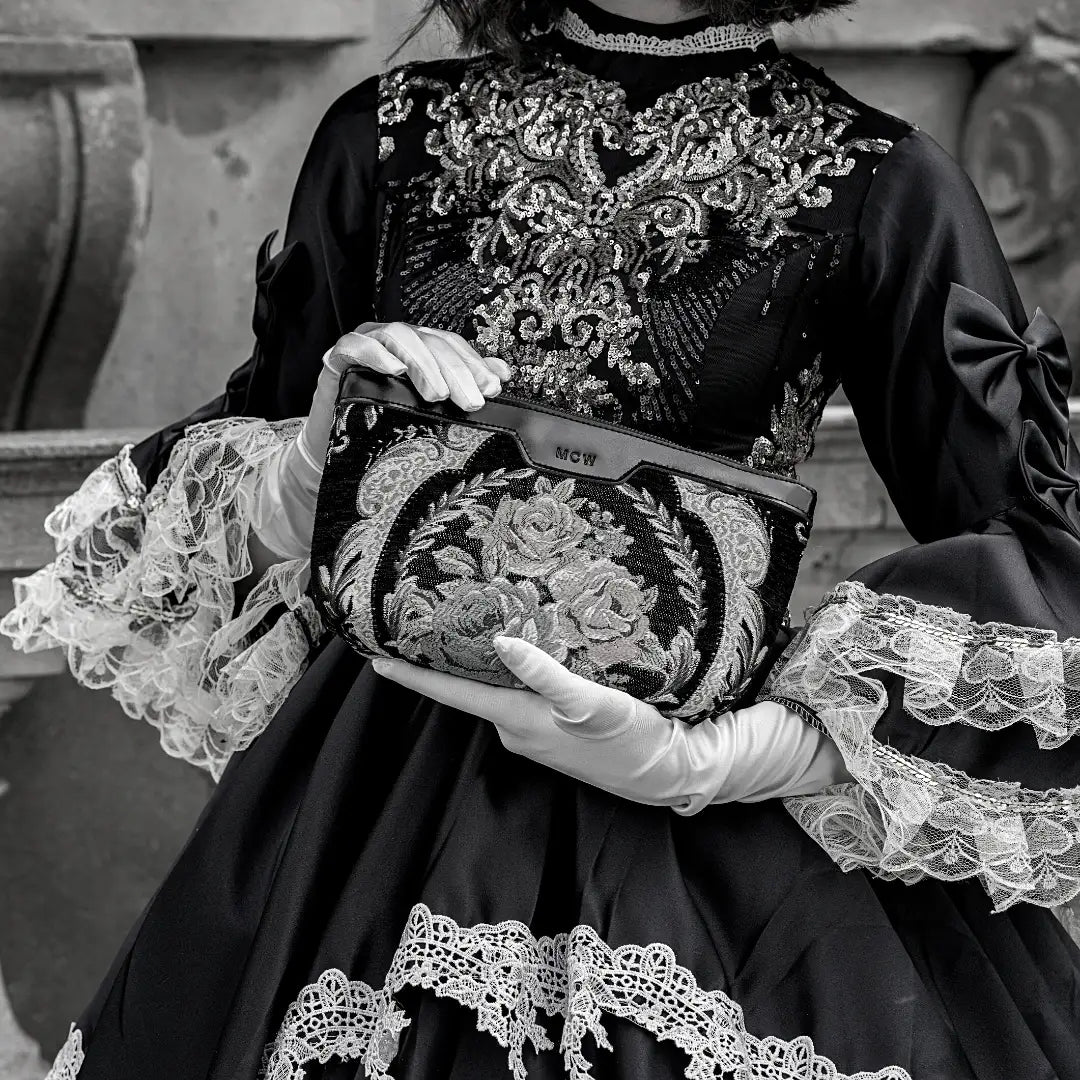 A black-and-white fashion portrait of a woman in vintage-style dress and wide-brim hat holding a patterned carpet clutch in front of a stone architectural background.
