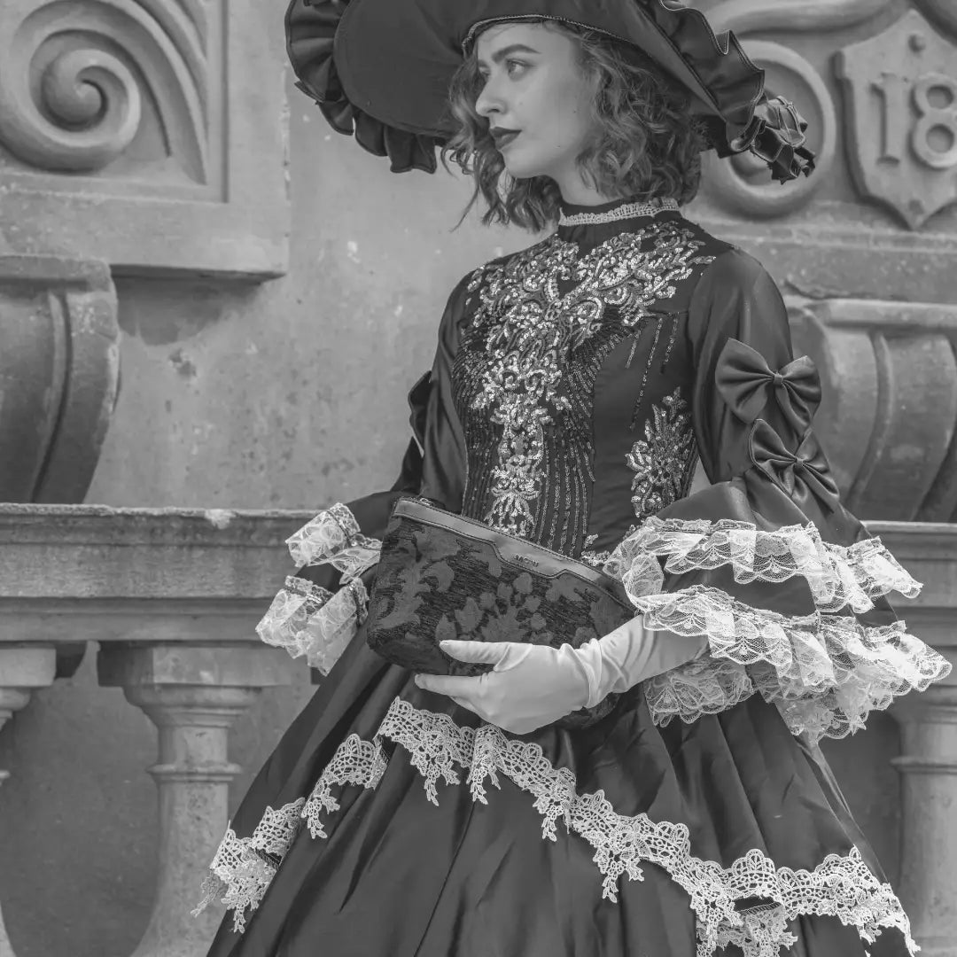 A black-and-white fashion portrait of a woman in vintage-style dress and wide-brim hat holding a patterned carpet clutch in front of a stone architectural background.