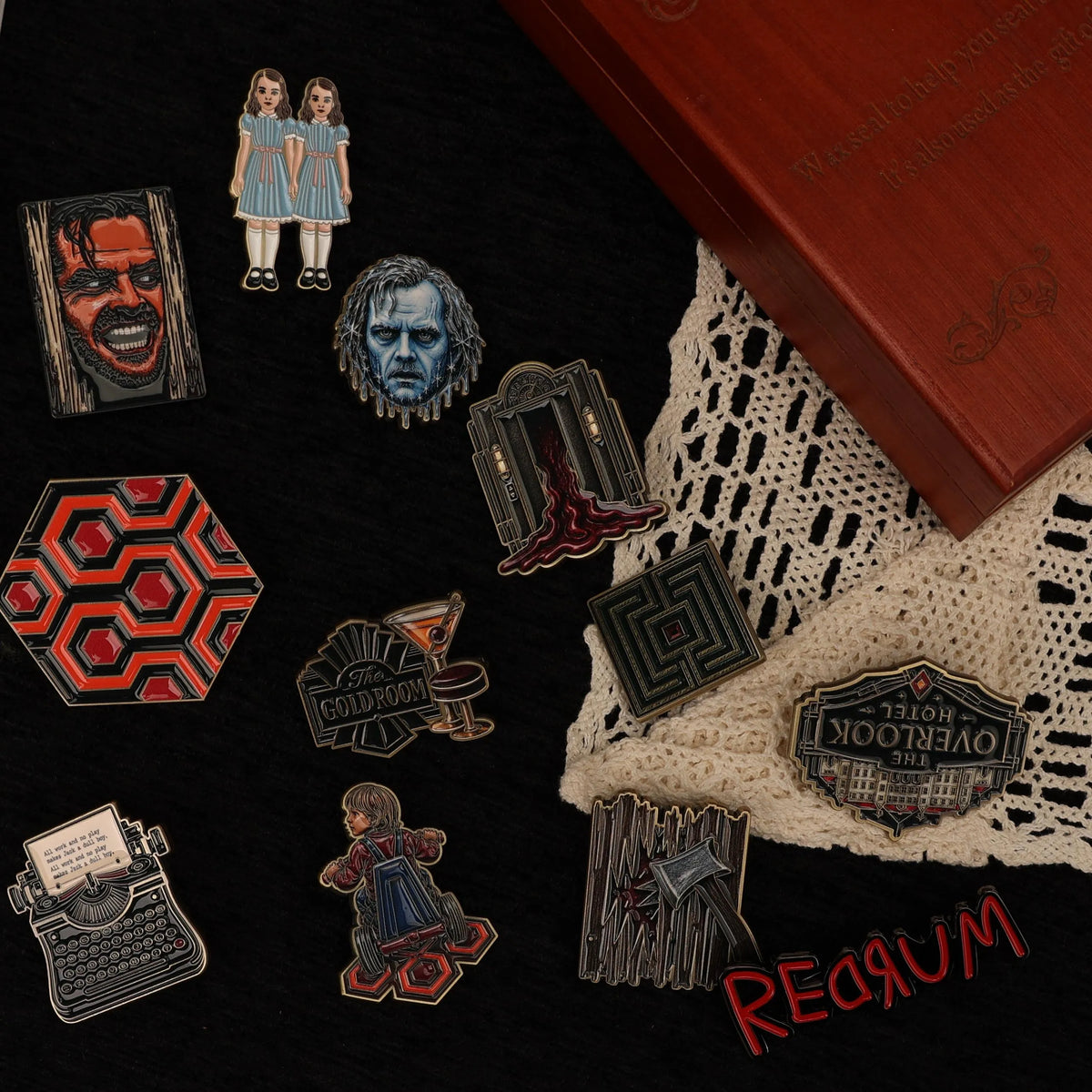 A set of horror-themed enamel pins arranged on a black background with a lace cloth, a brown box corner, and a red 'RED Rum' logo. The pins feature characters and symbols from The Addams Family / Wednesday series, including a portrait of Wednesday Addams, the Addams children, the Helix pattern, the Overlook Hotel sign, a typewriter, and other dark gothic motifs.