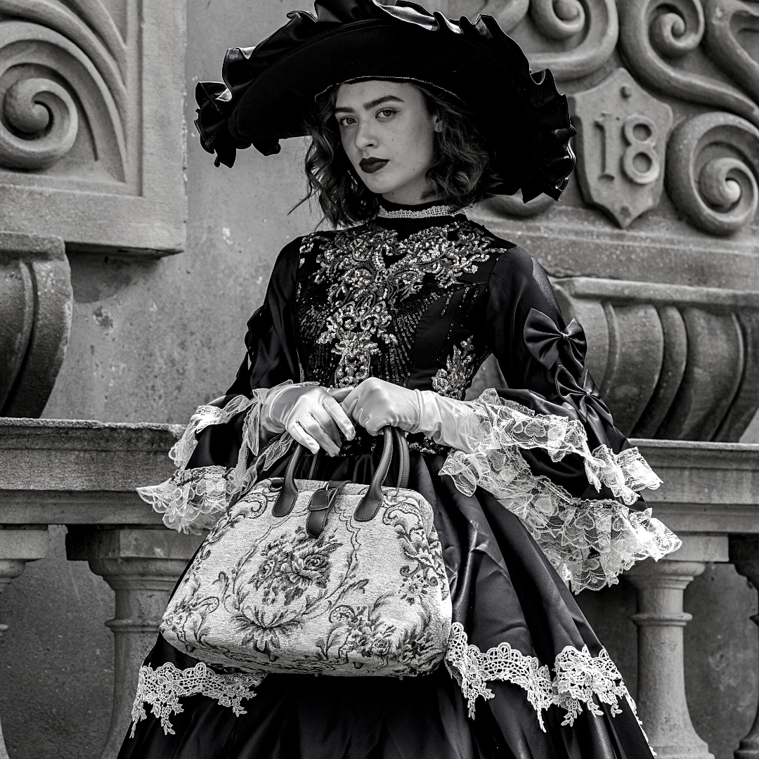 Woman in Victorian-inspired black lace dress and wide-brim hat holding a vintage floral carpet bag beside an ornate stone balcony in an elegant gothic setting.