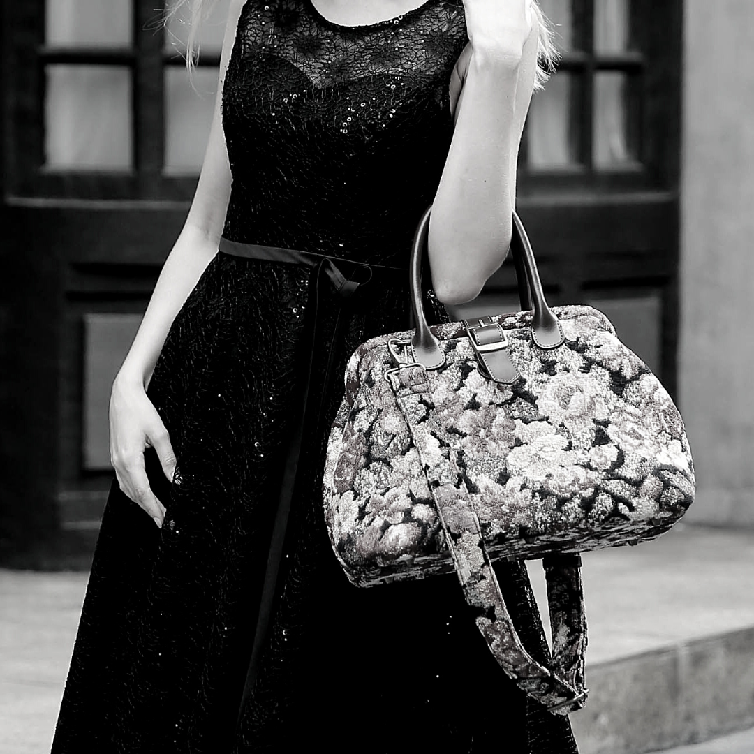 Woman wearing a black sequined dress carrying a vintage-inspired carpet handbag with leather handles and shoulder strap in an elegant city setting.