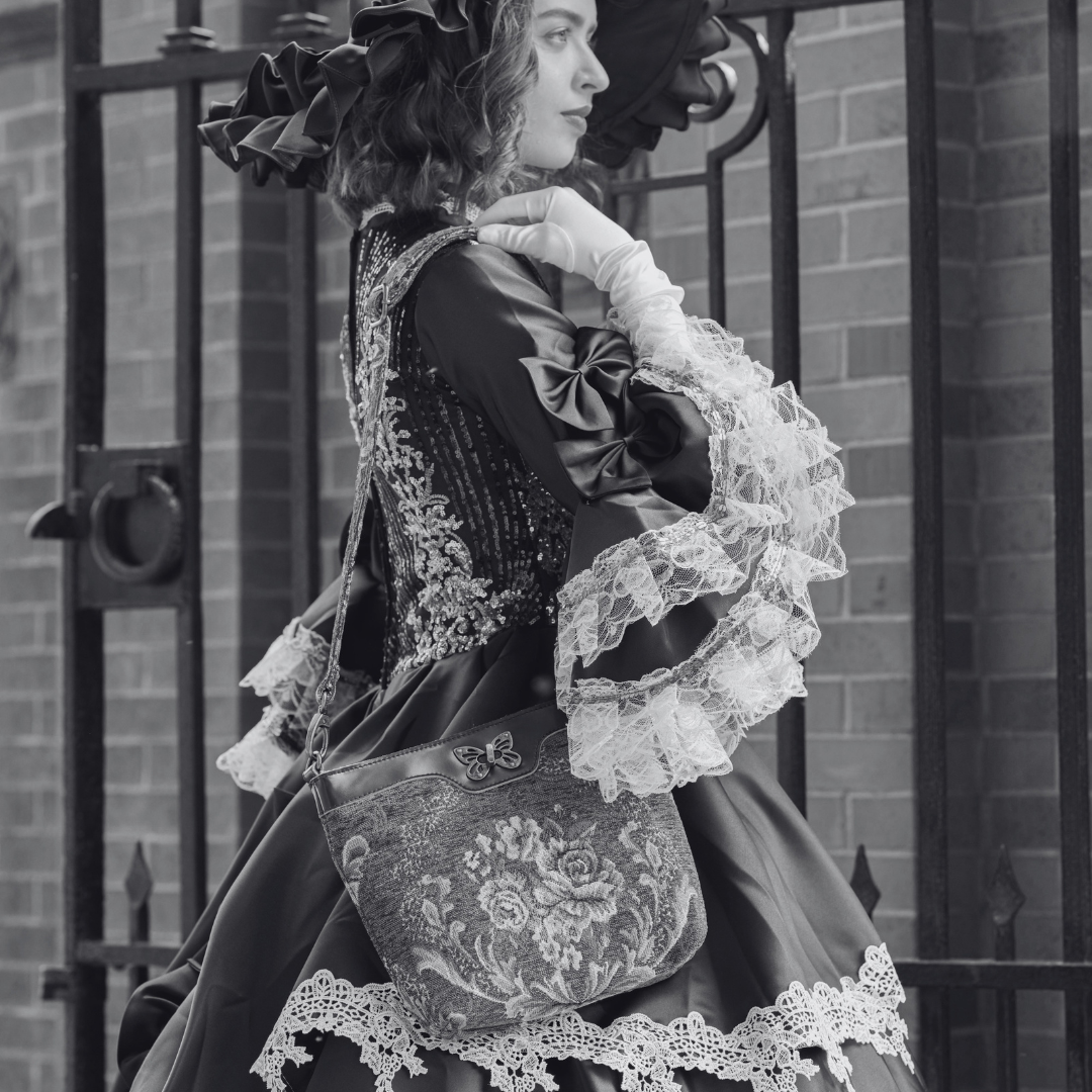 Woman in Victorian Gothic attire carrying a floral tapestry crossbody bag with a butterfly clasp beside an iron gate.