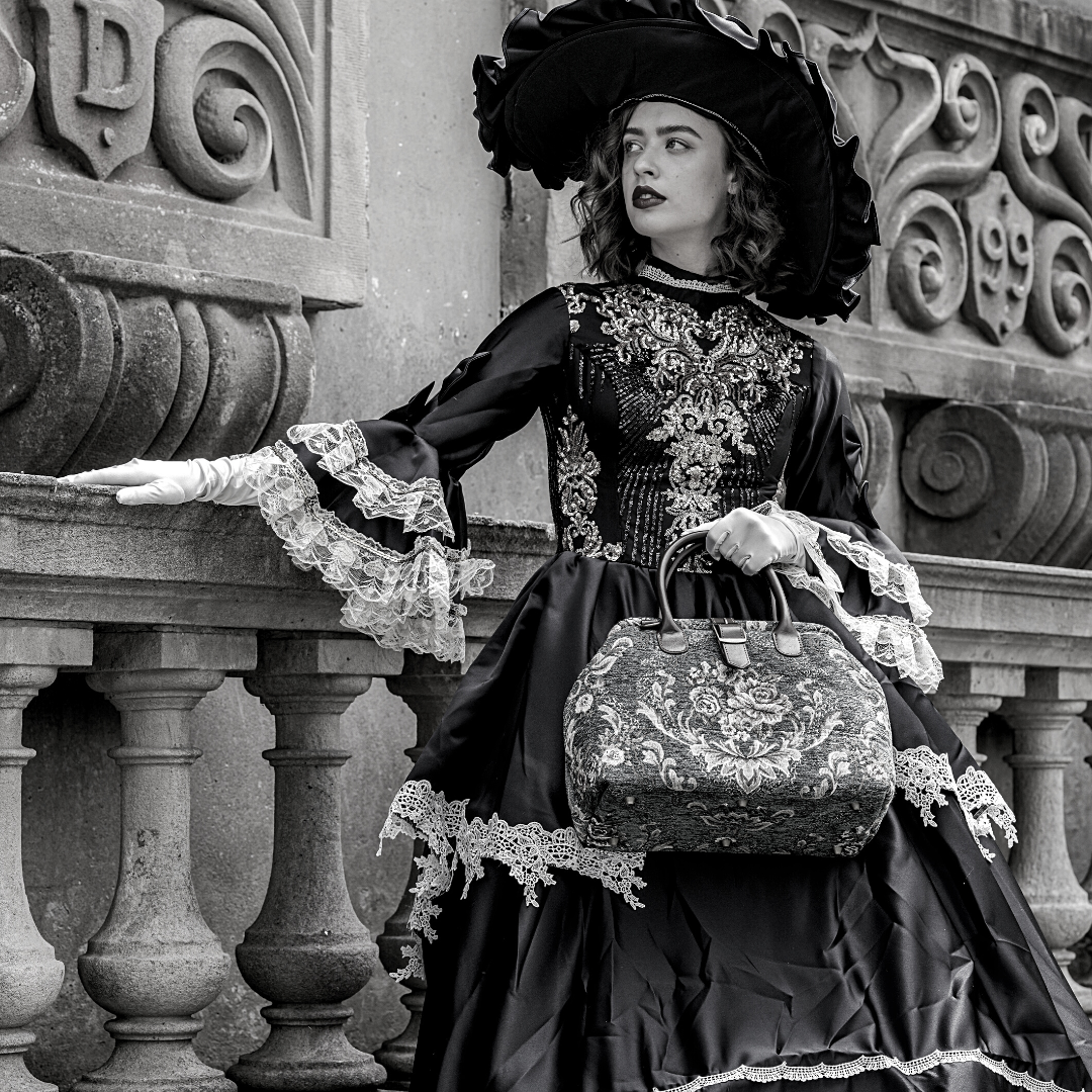 Woman in Victorian-inspired black lace dress and wide-brim hat holding a vintage floral carpet bag beside an ornate stone balcony in an elegant gothic setting.