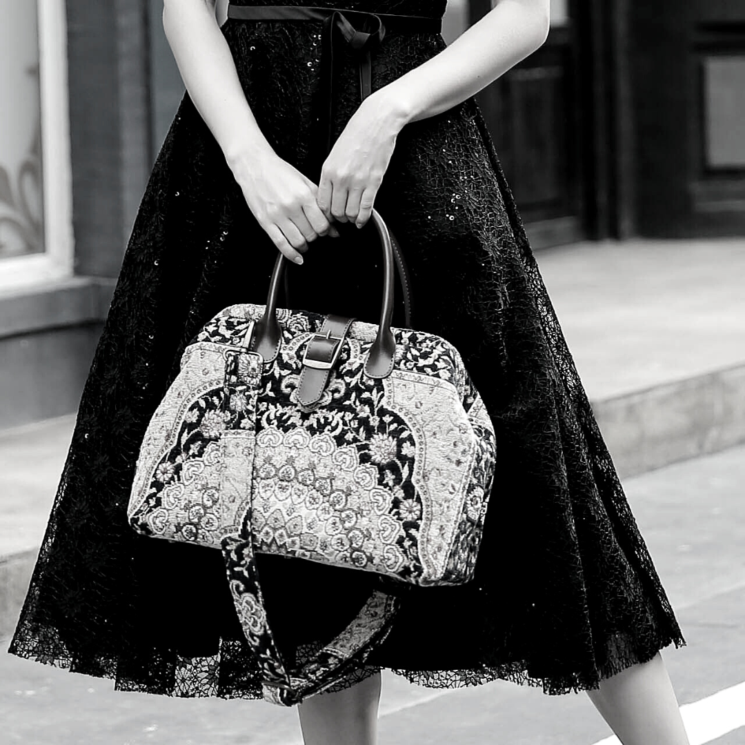 Woman wearing a black sequined dress carrying a vintage-inspired carpet handbag with leather handles and shoulder strap.
